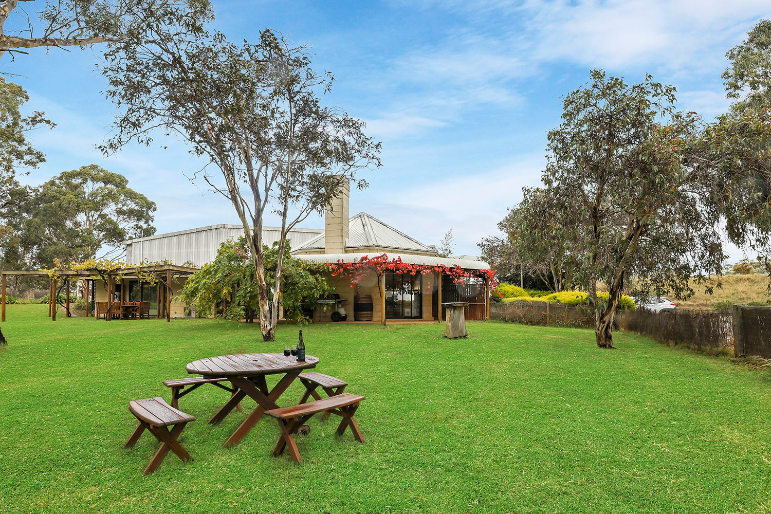 A group of wooden picnic tables arranged on a lush green lawn in front of a building with a bottle of wine and two glasses, with trees and the Mount Avoca cellar door building visible in the background.